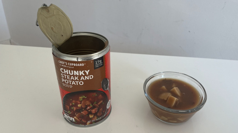 A can of steak and potato soup sits next to a glass bowl filled with it