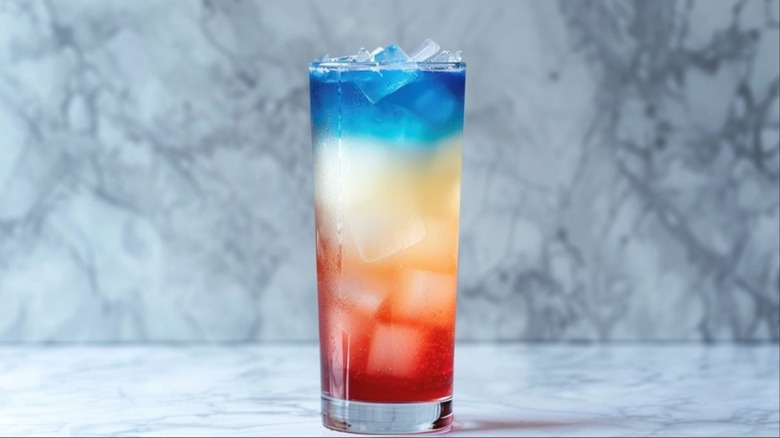 A red, white, and blue layered cocktail in a tall glass with ice cubes sitting on a kitchen counter
