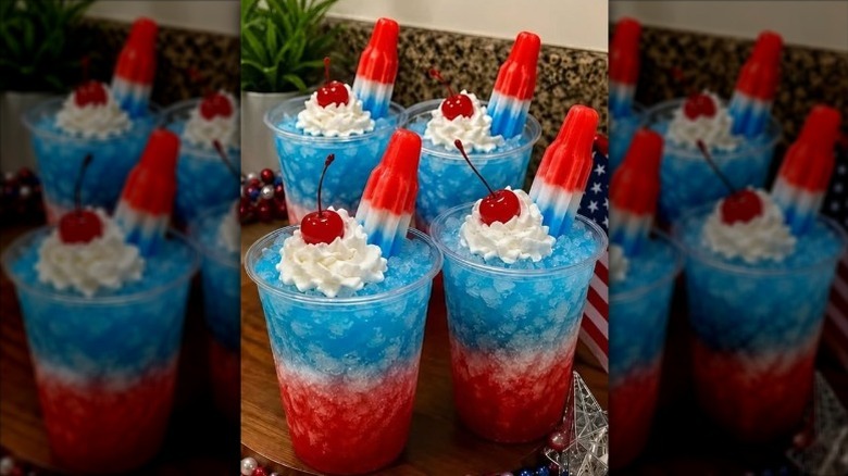 Four red, white, and blue layered cocktails in clear plastic cups of crushed ice garnished with whipped cream, maraschino cherries, and Bomb Pops