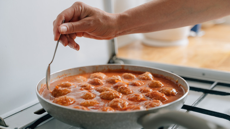 Meatballs simmering in pan on the stove with someone dipping a spoon into the sauce.