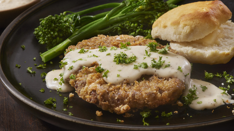 A piece of chicken-fried steak with white gravy on it beside a biscuit and tenderstem broccoli
