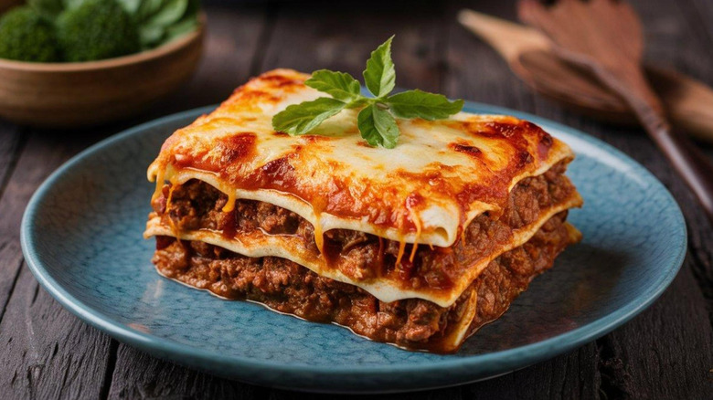 Square piece of meat-filled lasagna with melty cheese and scattered fresh herbs on a round plate