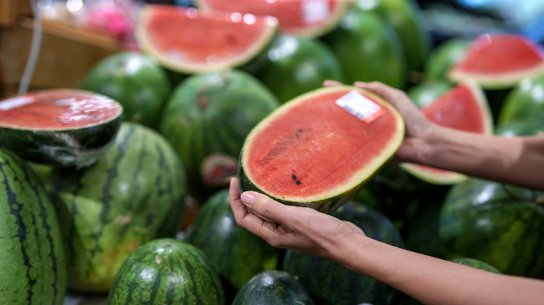 A woman's hands holding half a watermelon over a display of whole and cut watermelons