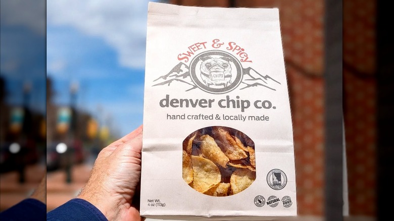 Hand holding brown paper bag of denver chip co. potato chips against brick building