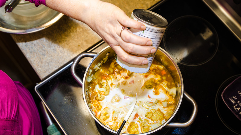 person pours can of coconut milk into soup