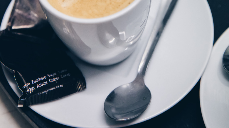 A white cup of coffee on a matching small plate with a black package of sugar sits to the left, a demitasse spoon to the right.