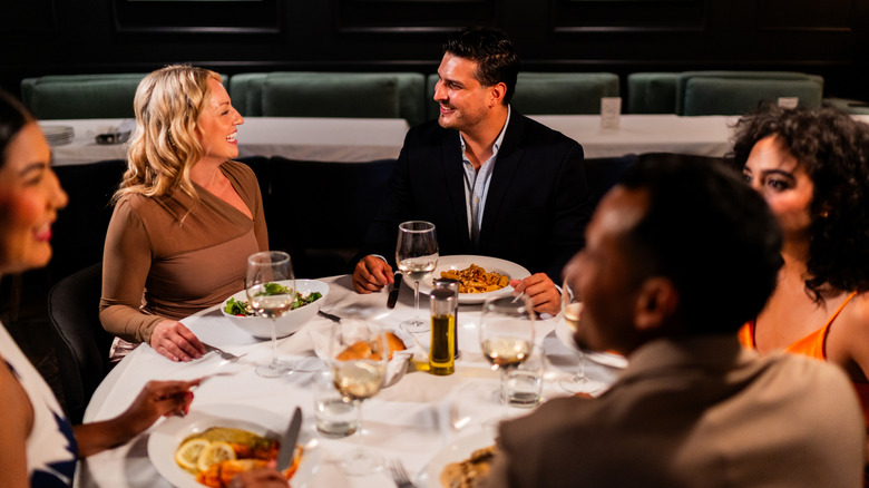 Five people sit around a table eating, drinking, talking, and laughing