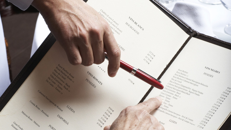 A waiter points to a menu item with his pen, the hand of the customer points underneith.