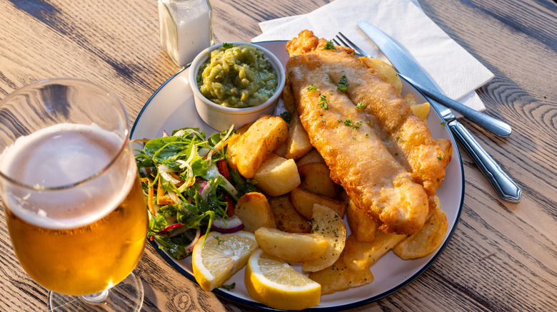 A plate of fish and chips served on a wooden table top, with mushy peas in a ramekin and a beer to the left of the plate