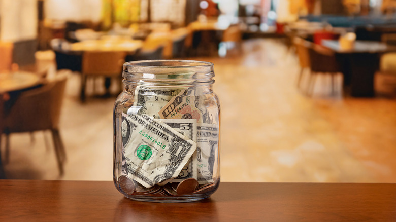 A glass jar filled with tips rests on a wooden table, a blurred out restaurant dining room in the background