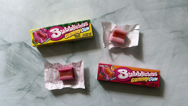 two packs of Bubblicious Gummy Gum next to two unwrapped pieces of the gum. One flavor is strawberry and orange and the other is watermelon.