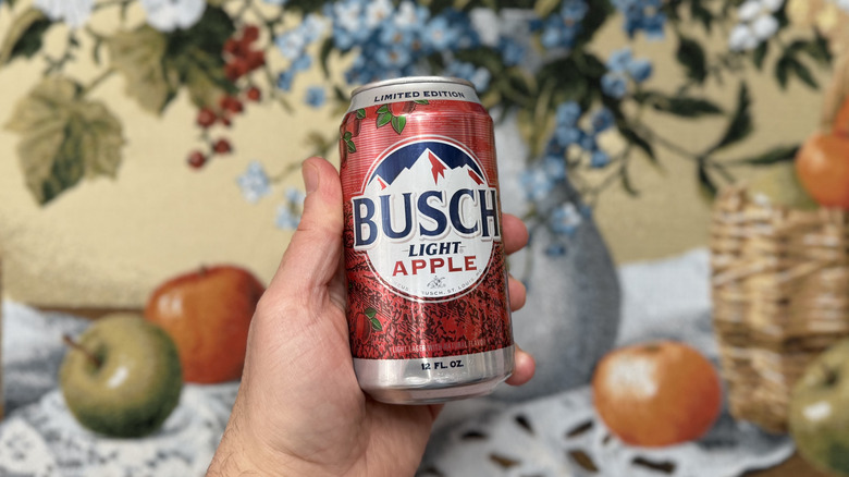 Can of Busch Light Apple held up against a tapestry of flower in vase and apples strewn on table
