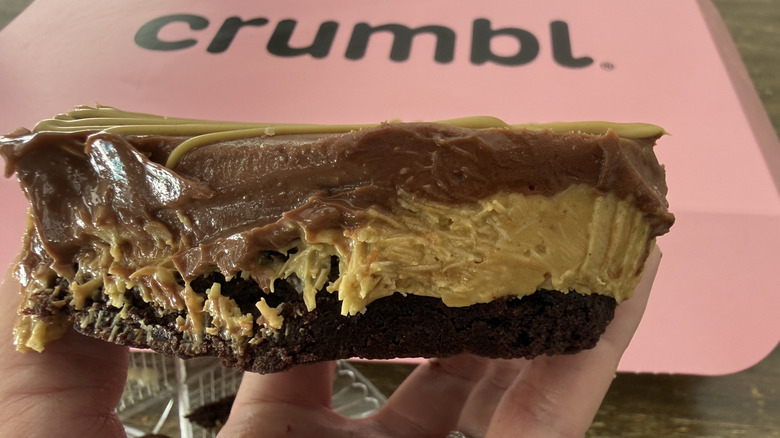 Hand holding a Dubai Brownie bitten in half to show the gooey inside with a pink Crumbl box in the background