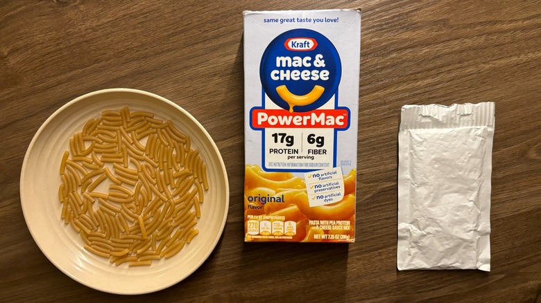 Kraft PowerMac box and dry noodles on a plate next to a white packet of Kraft Mac and Cheese powder mix