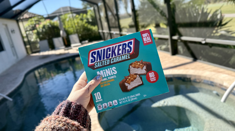 A hand holding a box of Snickers Salted Caramel Ice Cream Minis bars in front of a pool