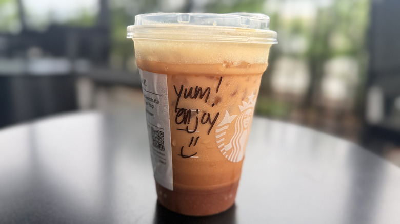 Writing on the side of a plastic Starbucks to-go cup sitting on a black table that reads "Yum! Enjoy" with a happy face