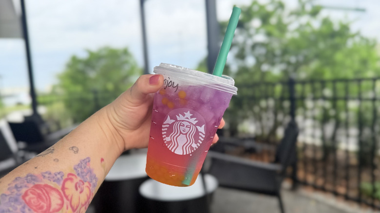 Hand holding a Tropical Butterfly Lemonade in a plastic to-go cup with a green straw in it