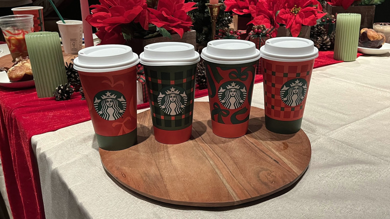 Four Starbucks holiday cups arranged in a row with a holiday-themed background