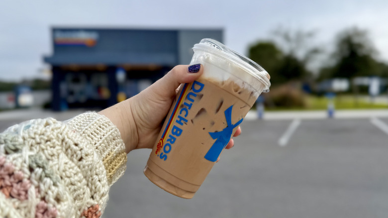 Close up of hand holding Dutch Bros Iced Banana Bread Mocha with Soft Top in parking lot in front of coffee shop