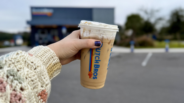 Close up of hand holding a Dutch Bros Iced Maple Waffle Latte in parking lot in front of coffee shop