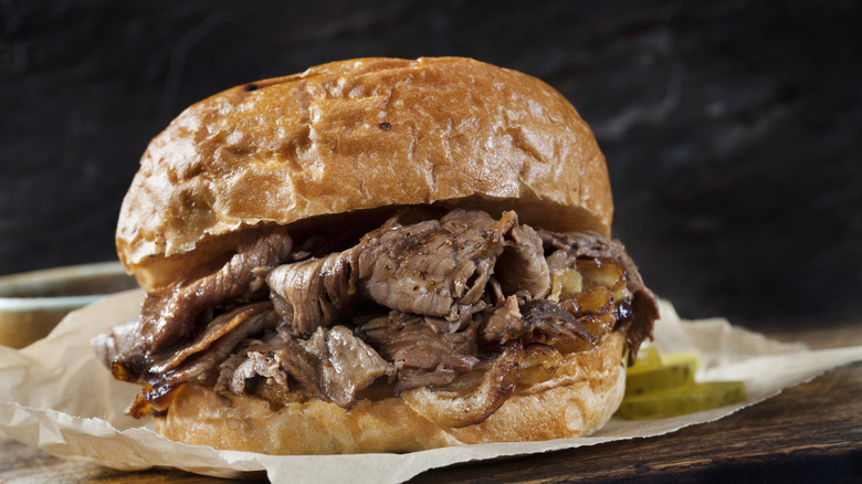 A roast beef sandwich sitting on brown paper with a side of pickles.