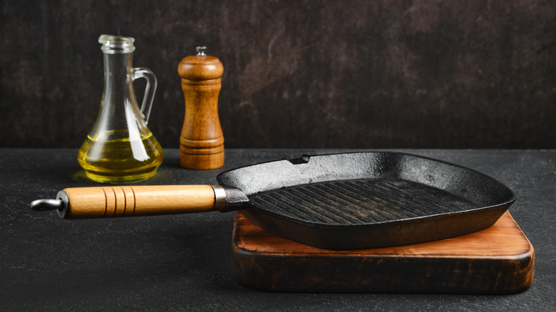 Square cast iron grill pan with wooden handle on a thick square cutting board with a wooden salt grinder and small oil jug
