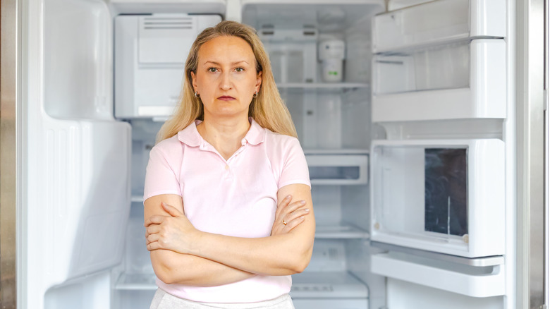 Woman standing in front of open, empty refrigerator with her arms crossed and blank look on her face