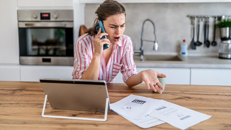 Woman in the kitchen arguing on the phone while looking at papers with a tablet nearby.