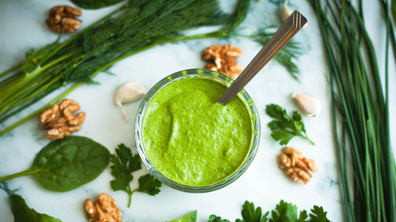 Thick, creamy green goddess dressing in a glass jar with a spoon, fresh herbs, and walnuts