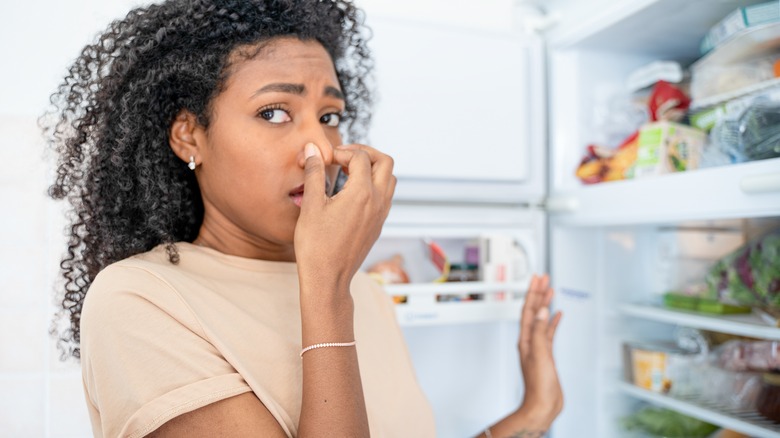 Woman holding nose in front of open refrigerator door