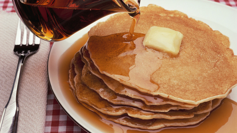 Pouring syrup on a stack of pancakes with a pat of butter on a white plate