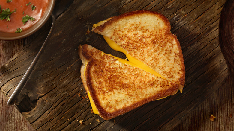 Sliced grilled cheese sandwich on dark wood cutting board next to bowl of tomato soup