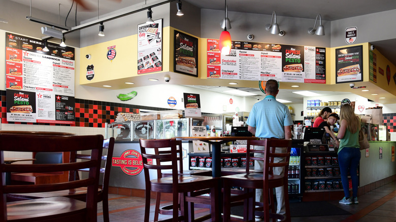 Interior of a Jimmy John's menus covering the walls and people standing at the counter placing an order.