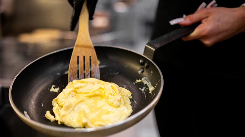 A chef holds a wooden spatula and a pan with scrambled eggs inside.