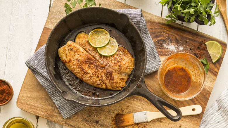 A seasoned tilapia filet sitting in cast iron pan with slices of lime.