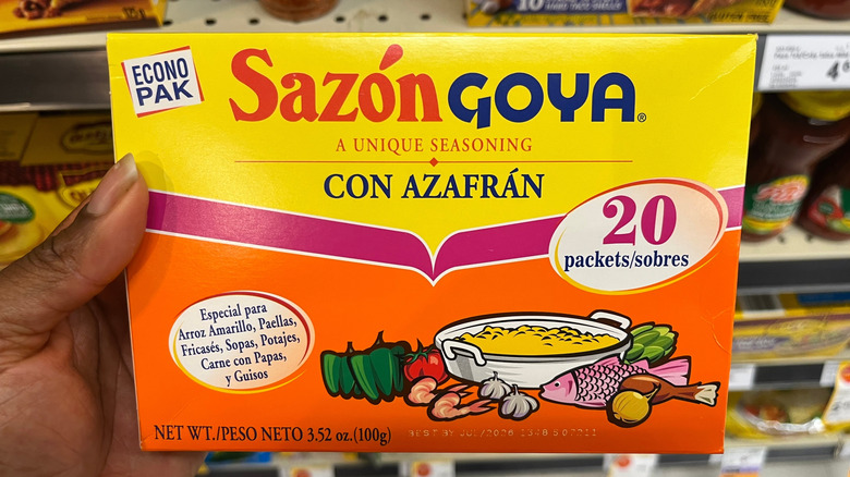 A person holding a box of Sazón Goya seasoning.
