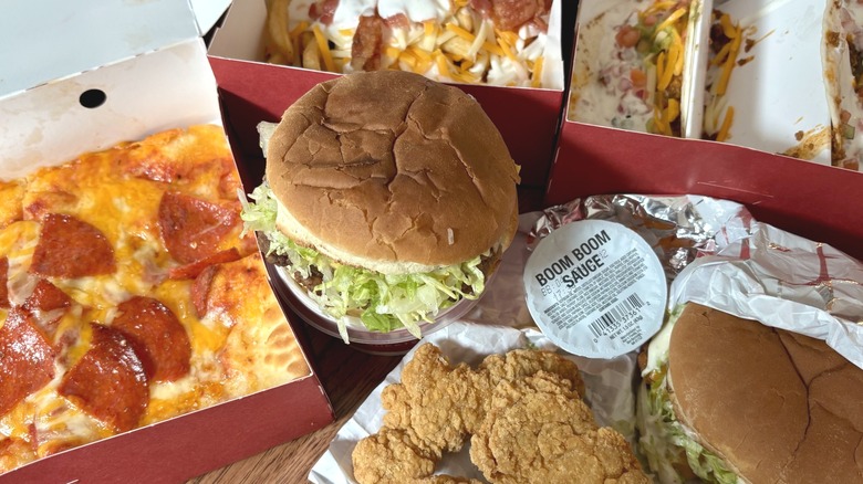 array of hot food options from Sheetz in takeout boxes on table, including pepperoni pizza, chicken sandwich, and hamburger
