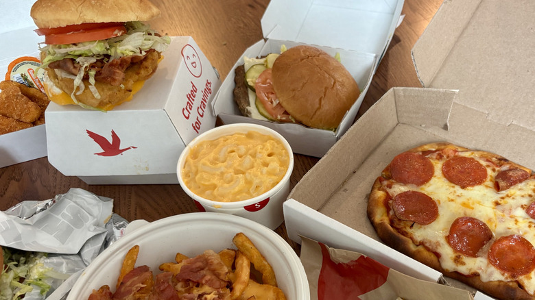 array of hot food from Wawa on table in takeout boxes, including cup of mac and cheese, pepperoni pizza, and burger