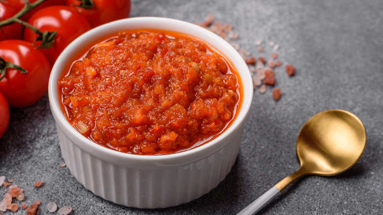Ramekin of tomato sauce with spoon and tomatoes nearby.