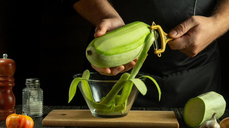 A person peeling raw zucchini over a glass bowl