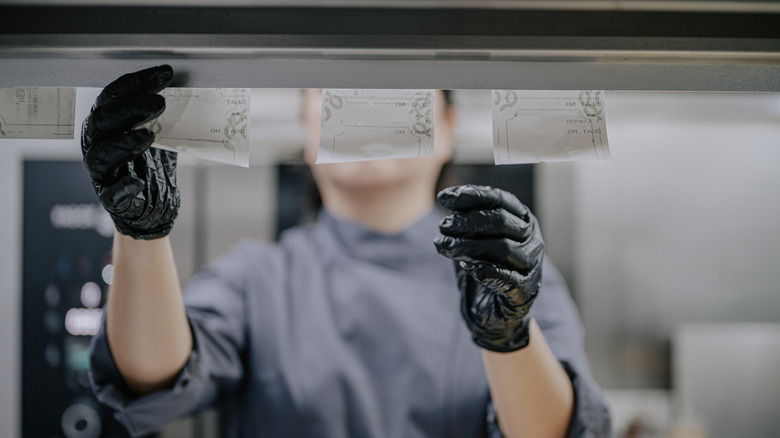 A chef looks at tickets above a kitchen pass, their face obscured by the tickets.