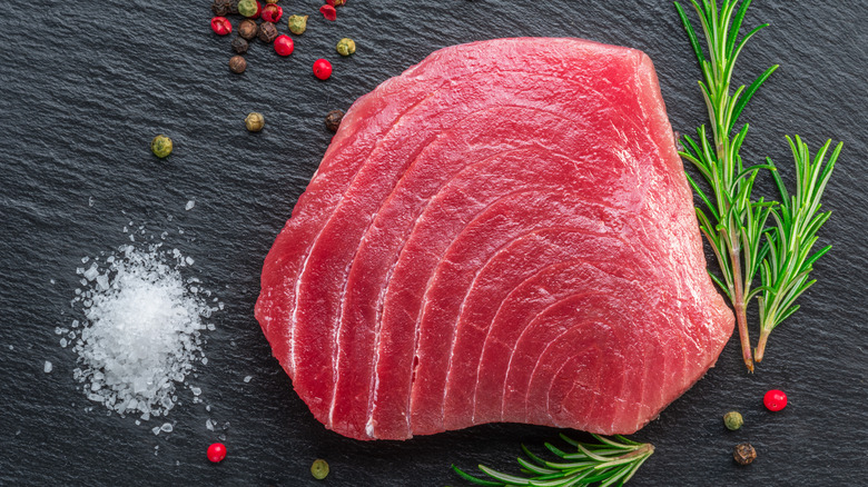 Raw tuna steak on stone surface surrounded by salt, peppercorns, and rosemary sprigs
