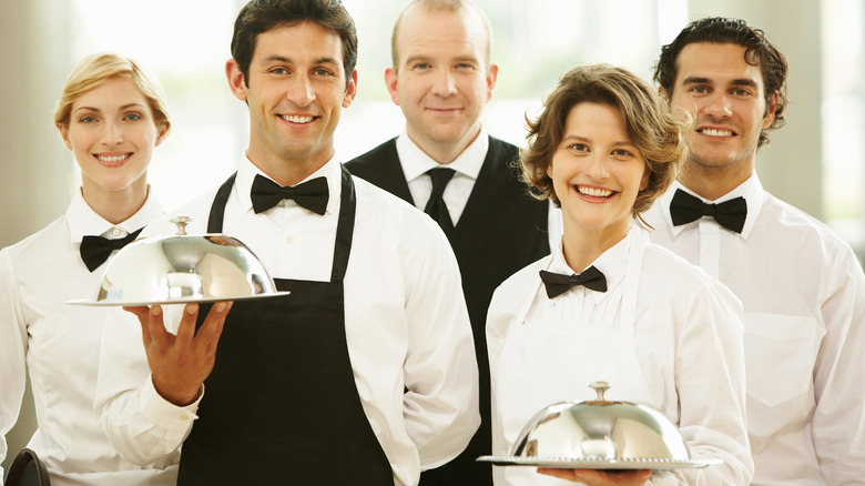 five servers dressed in uniforms and smiling at the camera, with two holding cloches