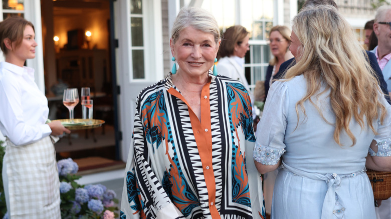 Martha Stewart stands outdoors at a garden party smiling surrounded by guests