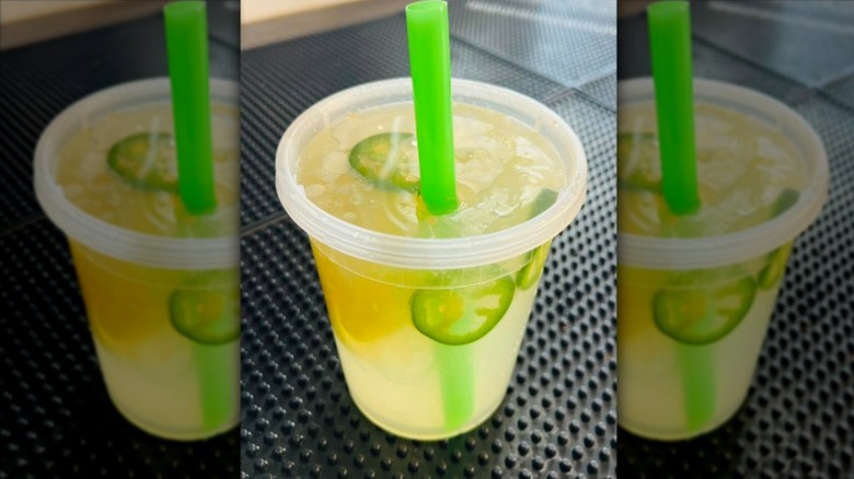 lemonade with floating slices of jalapeño and lemon in a lidded plastic cup with a green straw sitting on a textured black rubber mat