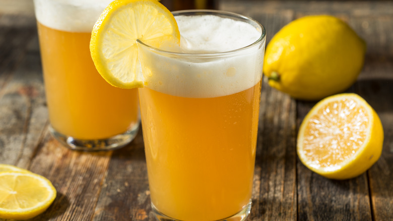 Shandy in a pint glass garnished with a lemon wheel sitting on a wooden table in front of another shandy and next to a whole and half lemon