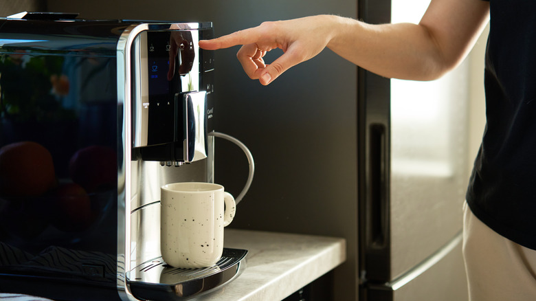 A woman pressing a button on a coffee maker machine with an empty mug under the spout.