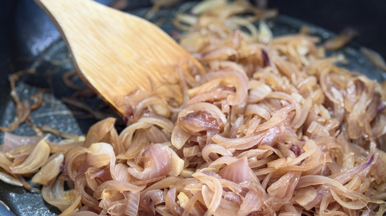 A wooden spoon stirs onion slices in a pan
