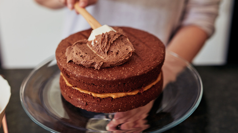 A baker in the process of frosting a chocolate cake on a turntable