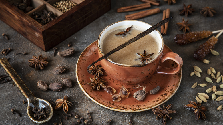A mug of chai in a terra cotta colored mug and saucer, surrounded by whole spices like cardamom, cinnamon, and star anise